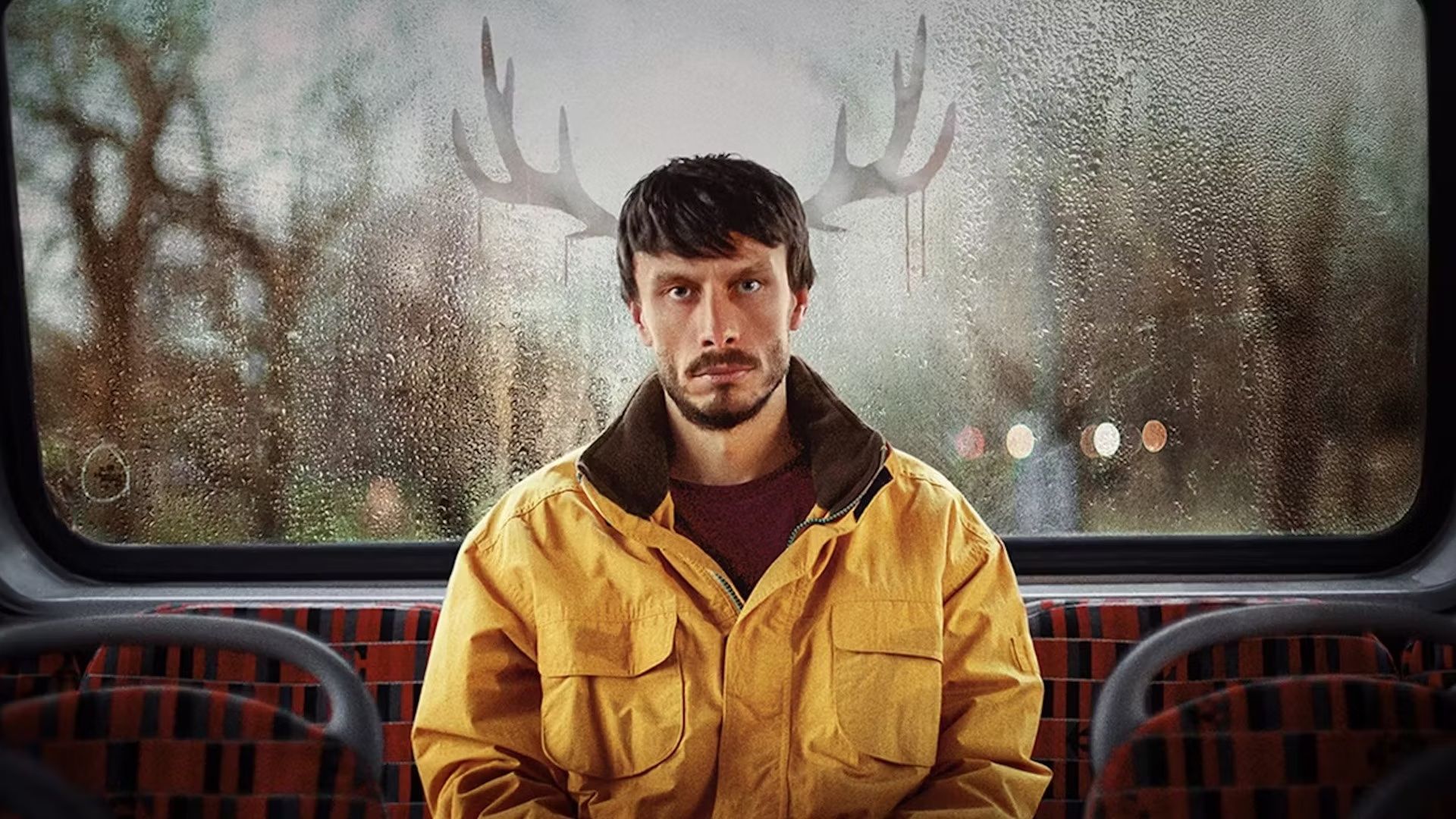 Donny sitting at the back of a bus with a yellow jacket, outline of reindeer ears on either side of him in the window in a scene from Baby Reindeer.
