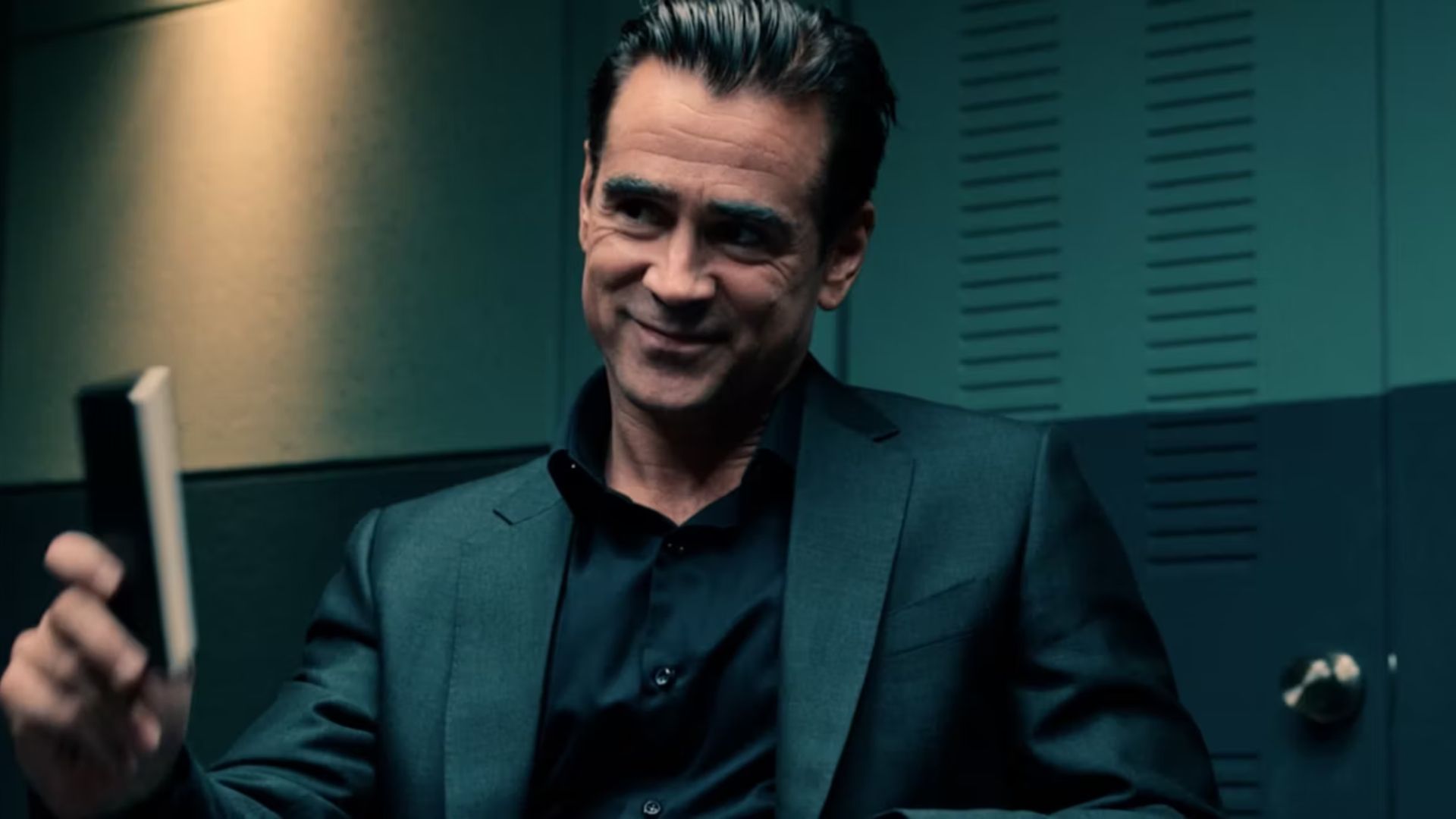 Colin Farrell as John Sugar holding a notebook sitting down wearing a black suit in Sugar