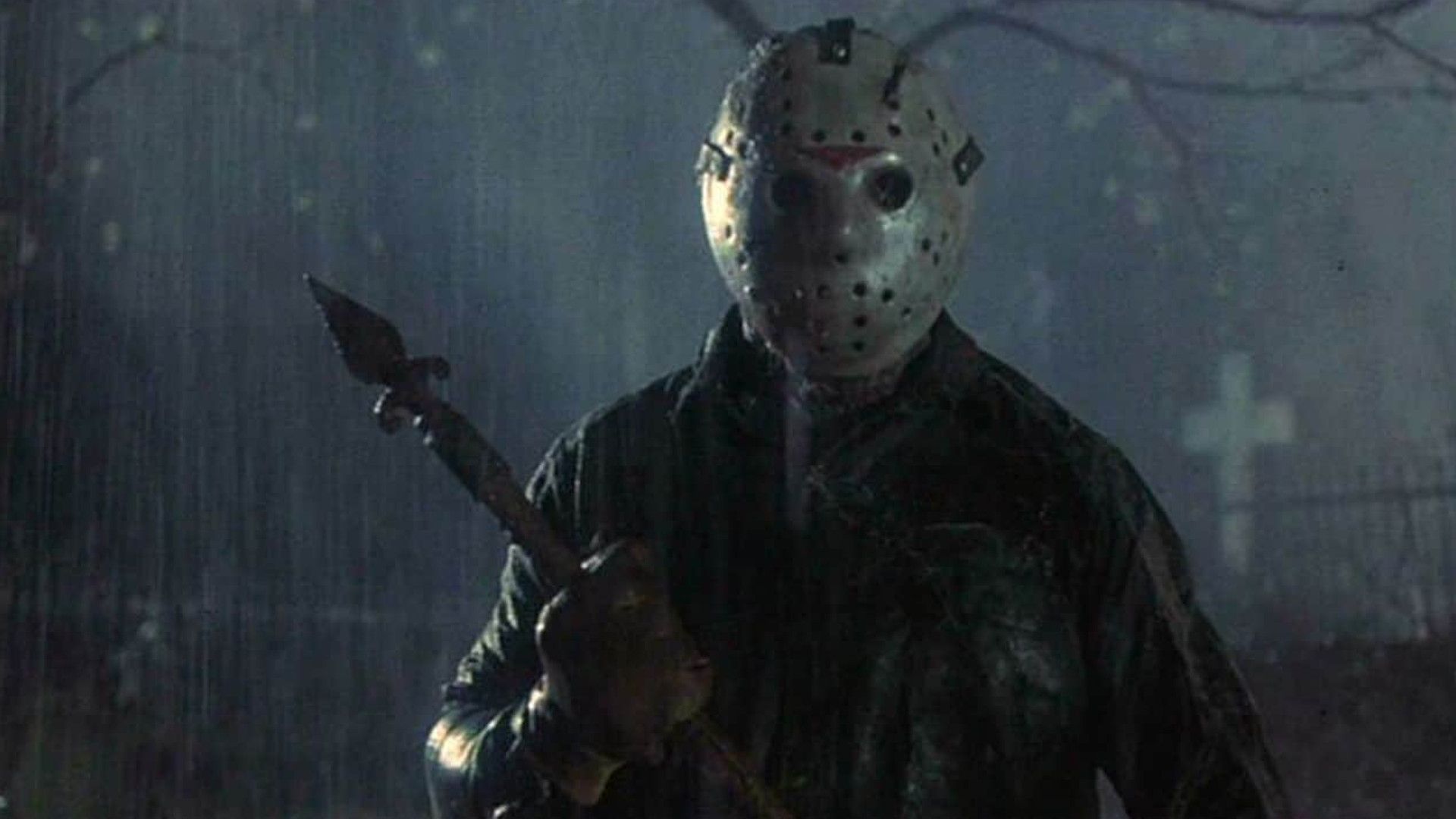 Jason Voorhees in Friday the 13th Part 6 in a graveyard wearing a hockey mask and holding a spear in the rain.