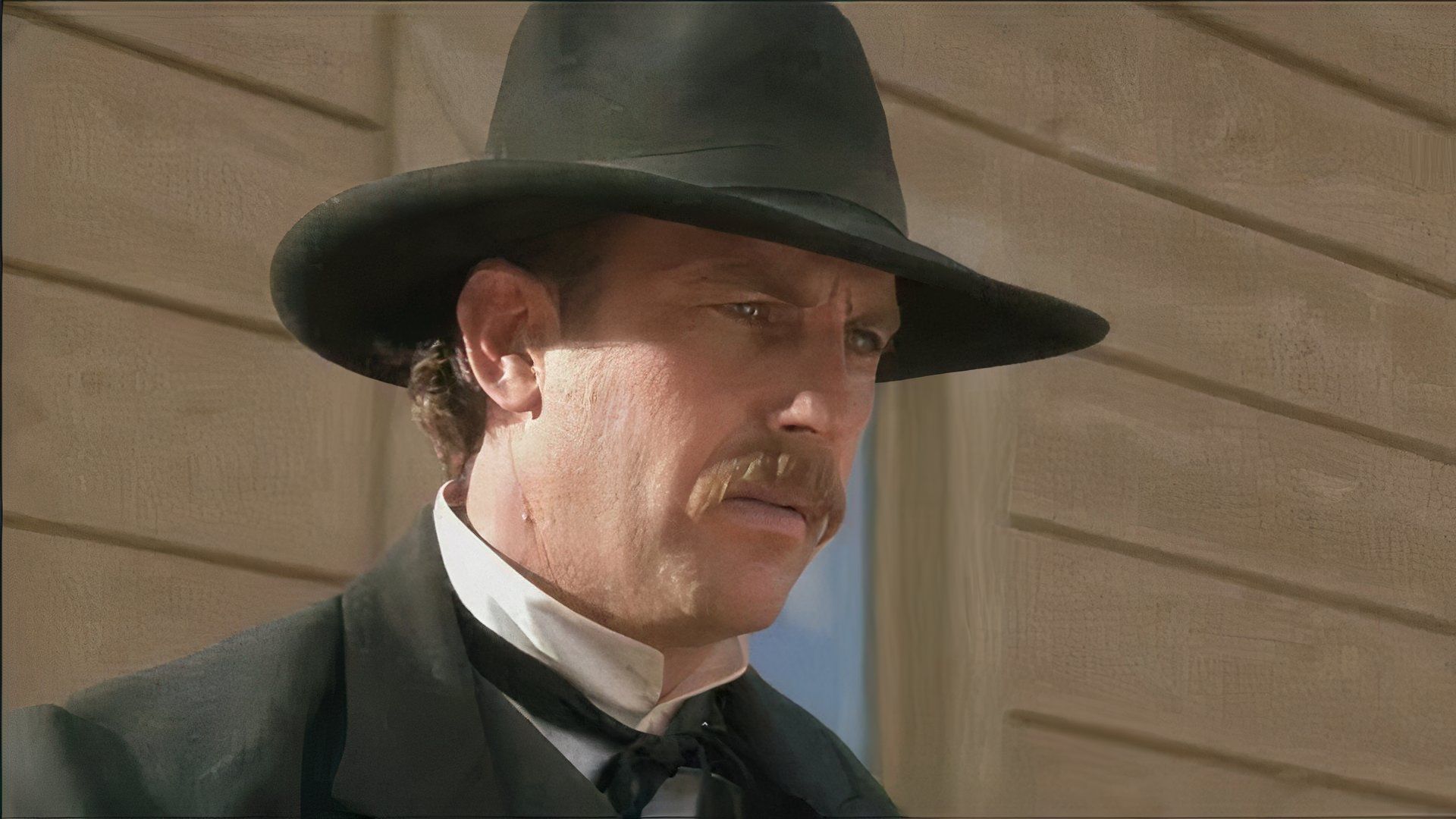 Kevin Costner wearing a black hat and black jacket looking off-screen in Wyatt Earp