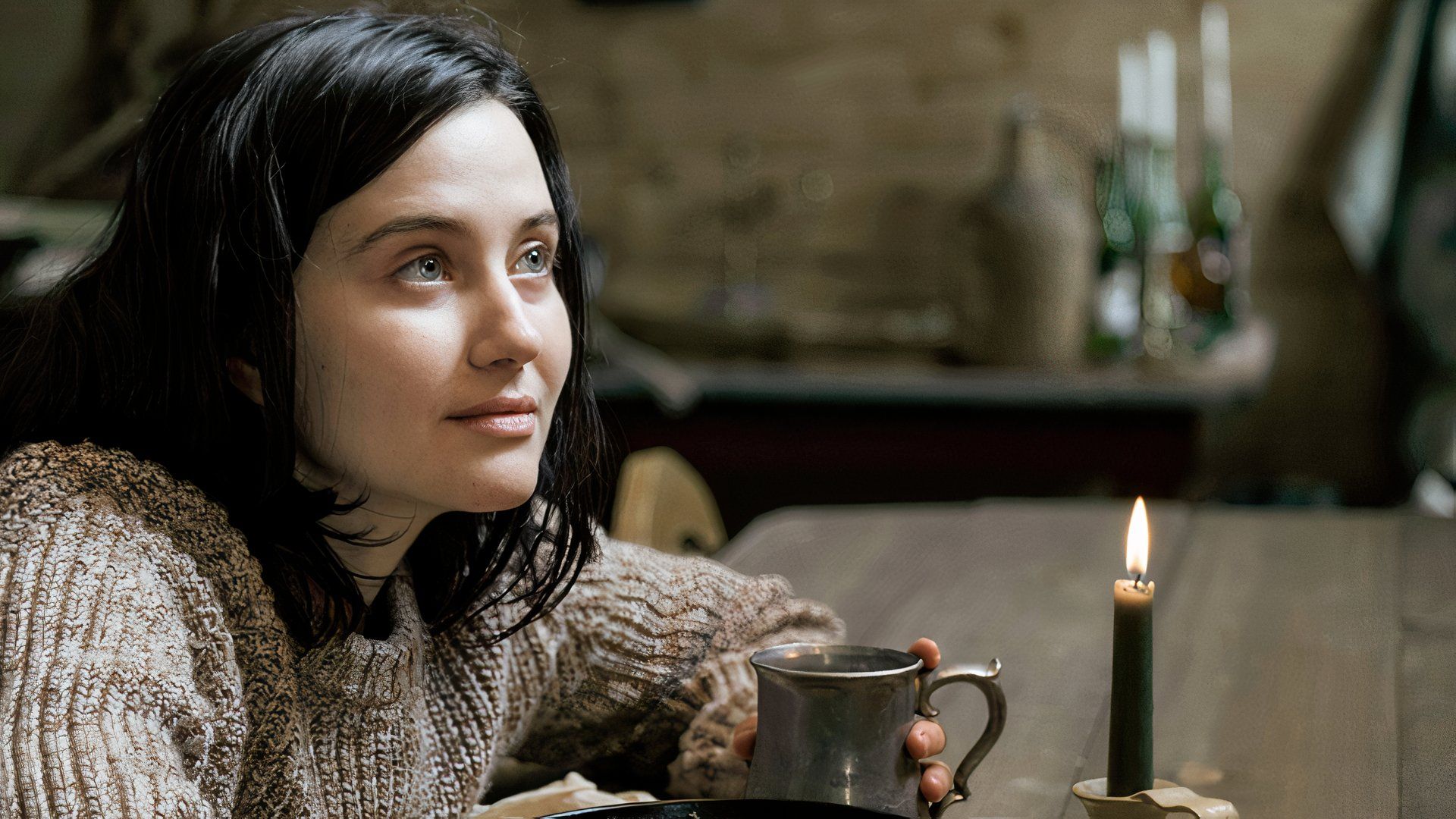 Julia Goldani Telles as Emily, sitting in a candlelit cabin and wearing a knitted sweater, in Beacon