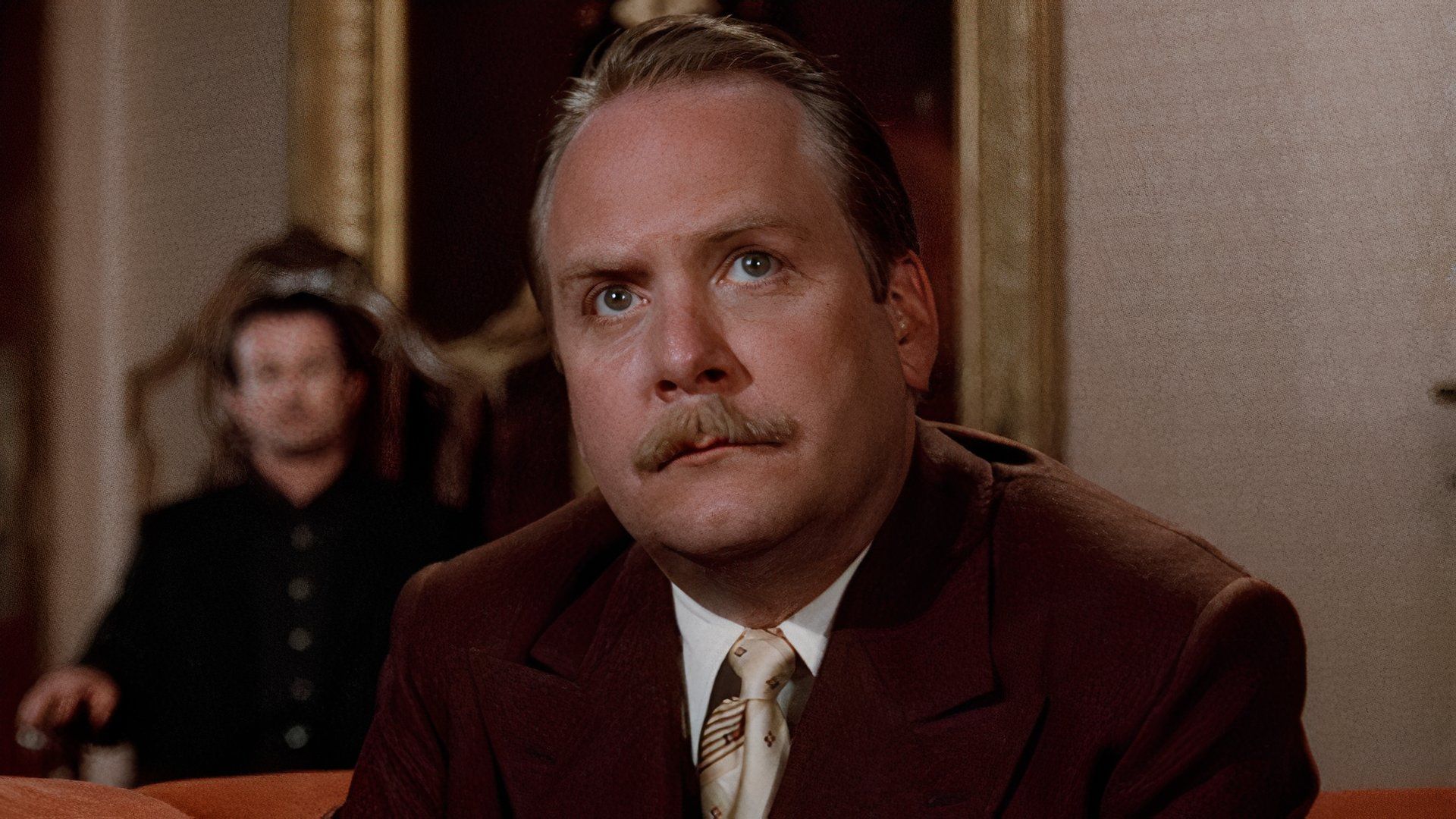 Martin Mull as Colonel Mustard sits before Mr. Body in Clue