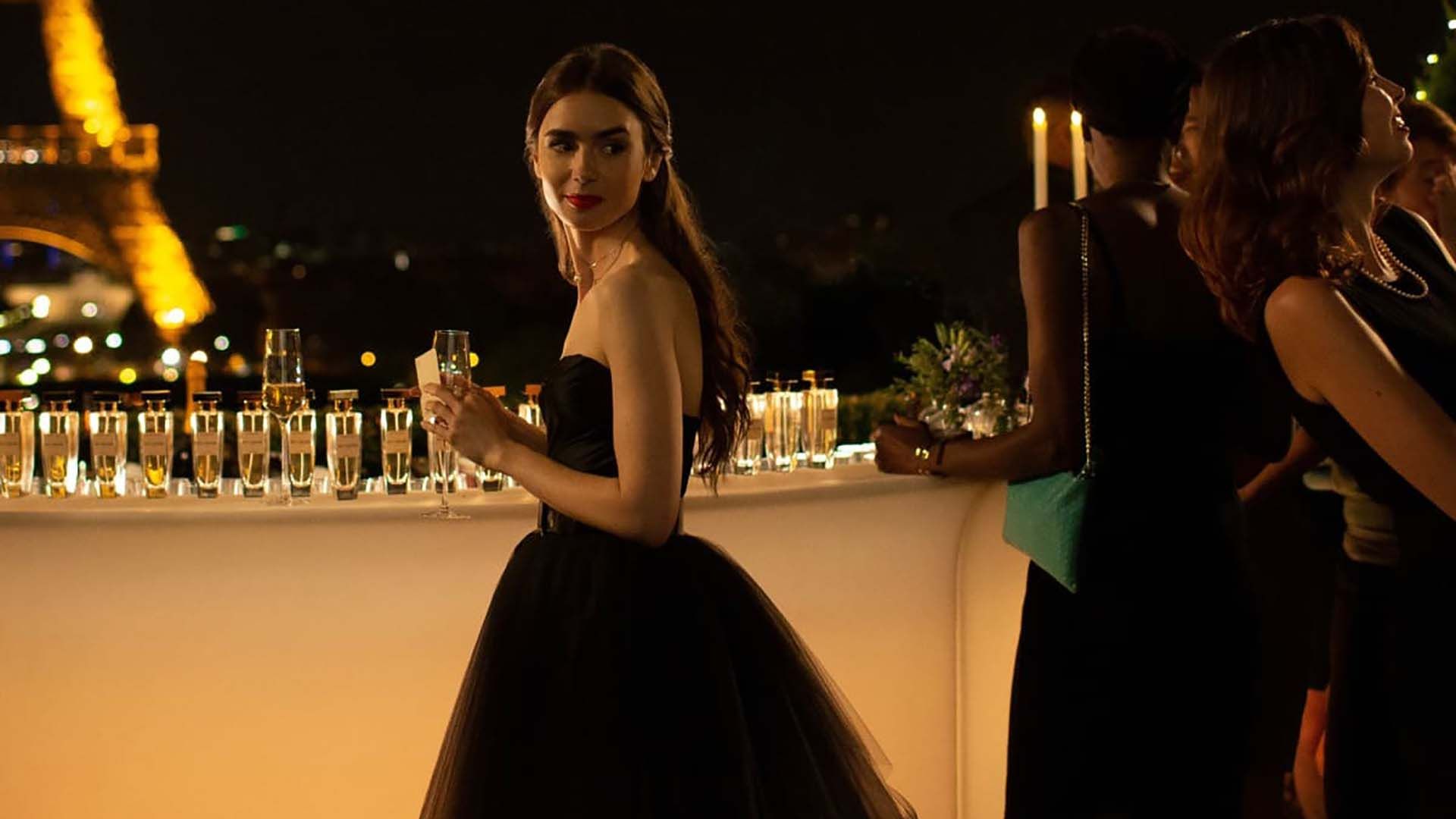 Emily on a rooftop in a black dress holding a drink and looking back on Emily in Paris.