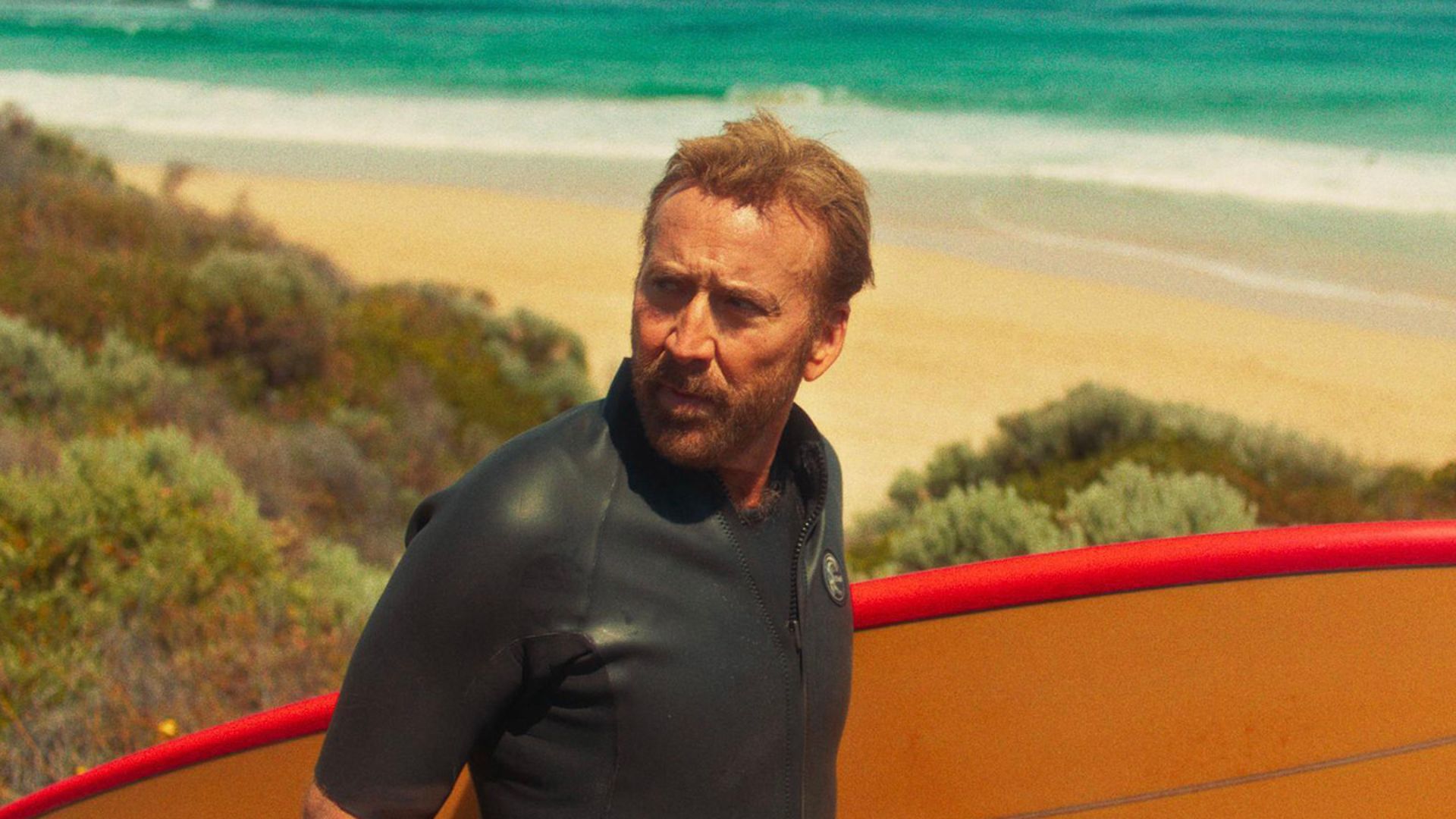 Nicolas Cage with a surfboard by the beach in the movie The Surfer