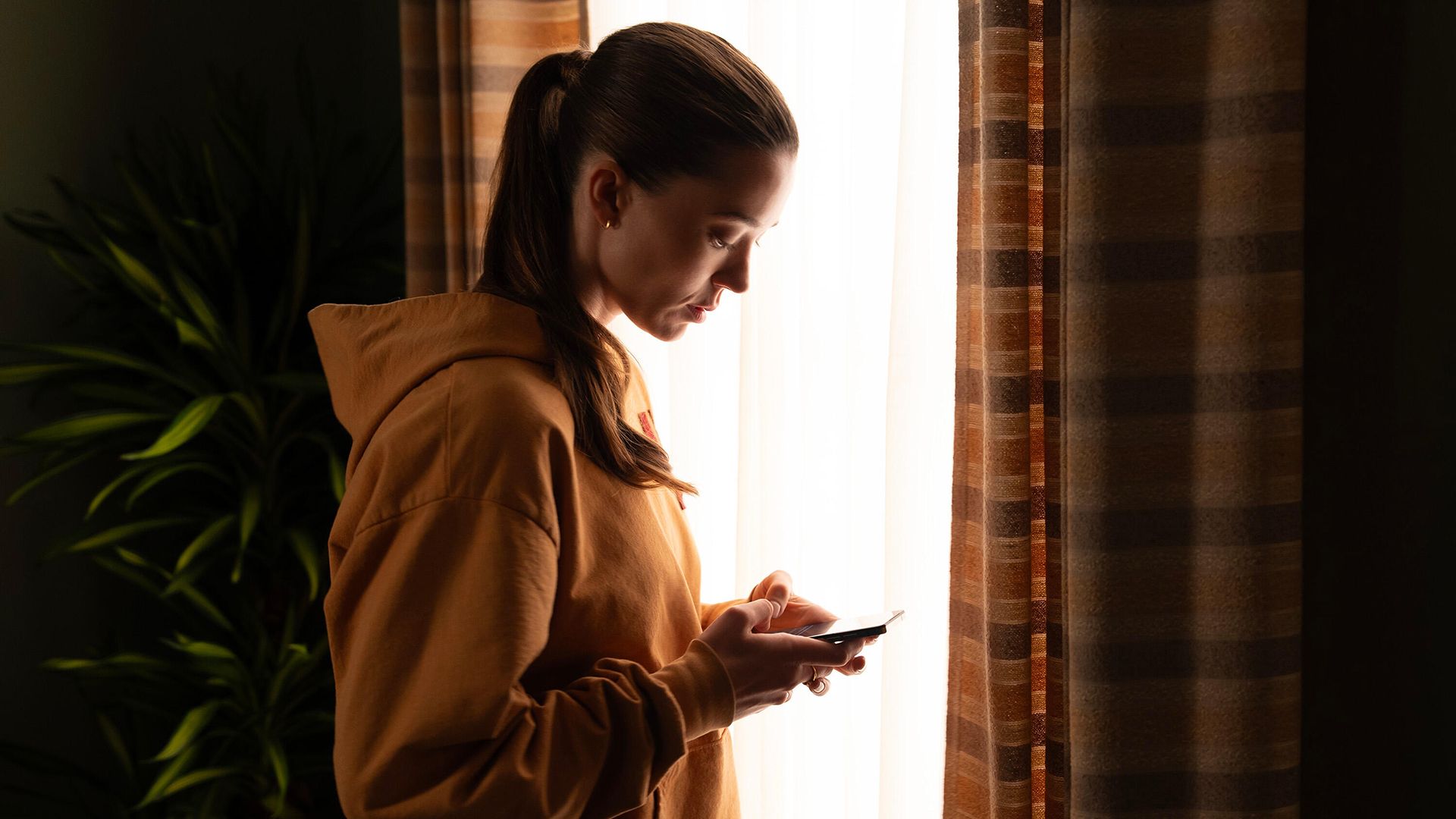 Jane standing by the window in a sweater on her phone in Paradise.