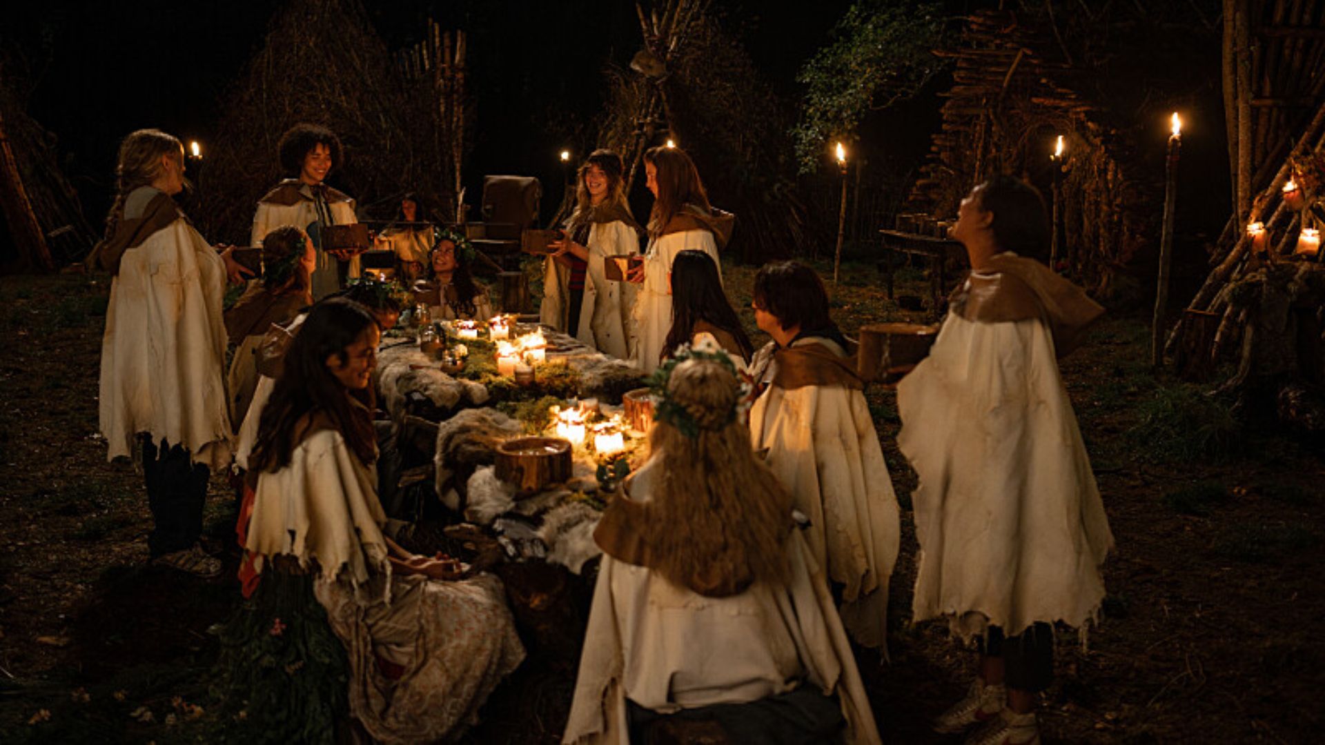 The teen cast in the wilderness in Yellowjackets in white cloaks sitting around a table