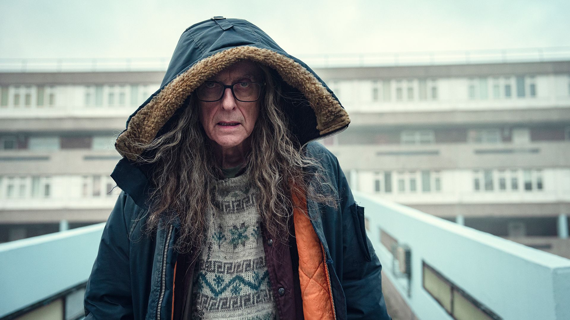 A man walking along a bridge with long hair and the hood of his jacket over his head in Black Mirror.