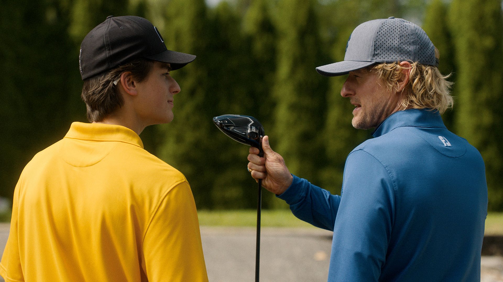 The backs of Santi and Pryce as Pryce hands him a golf club in Stick