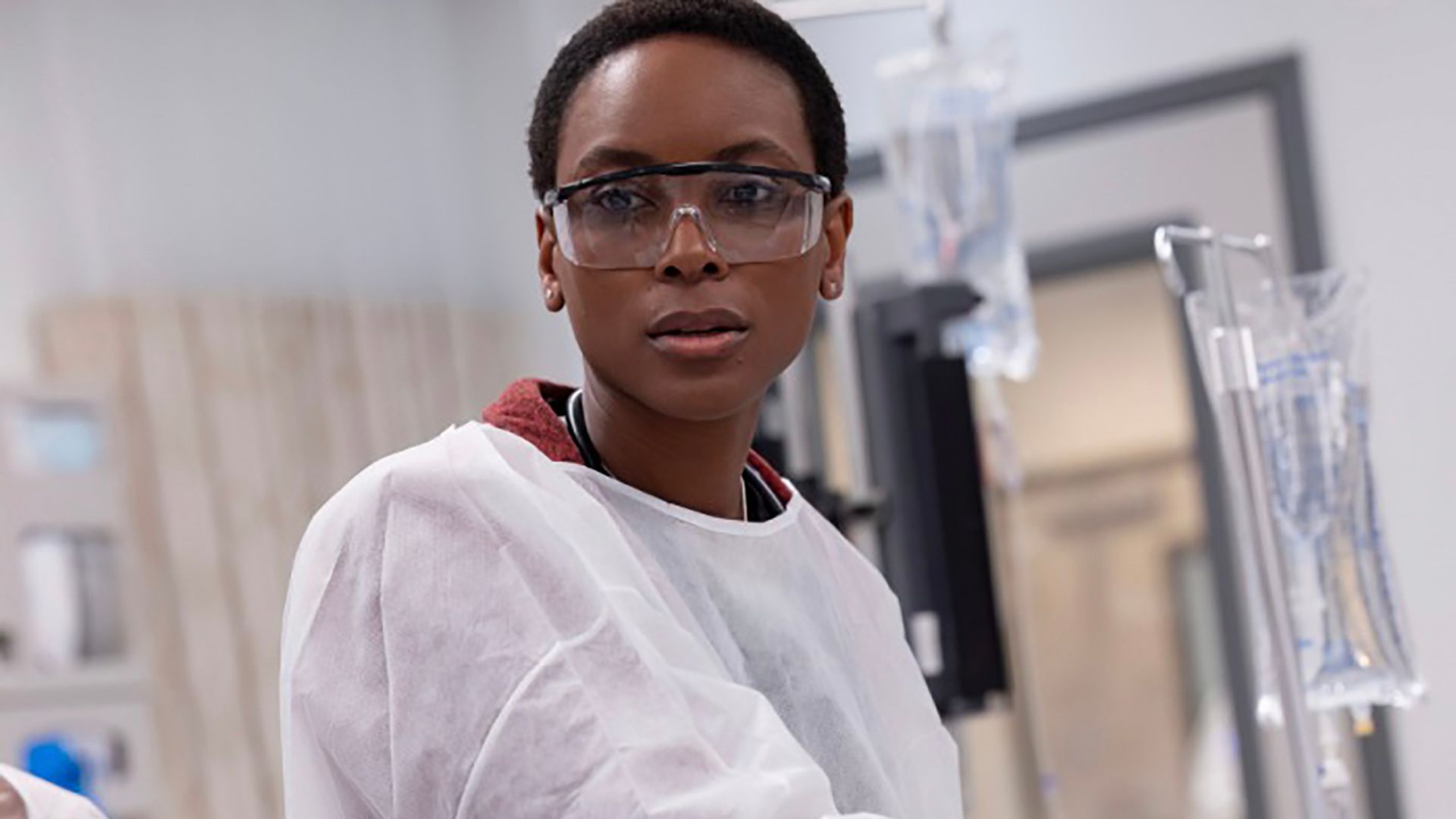 Dr. Collins with a white hospital gown and glasses in The Pitt. 