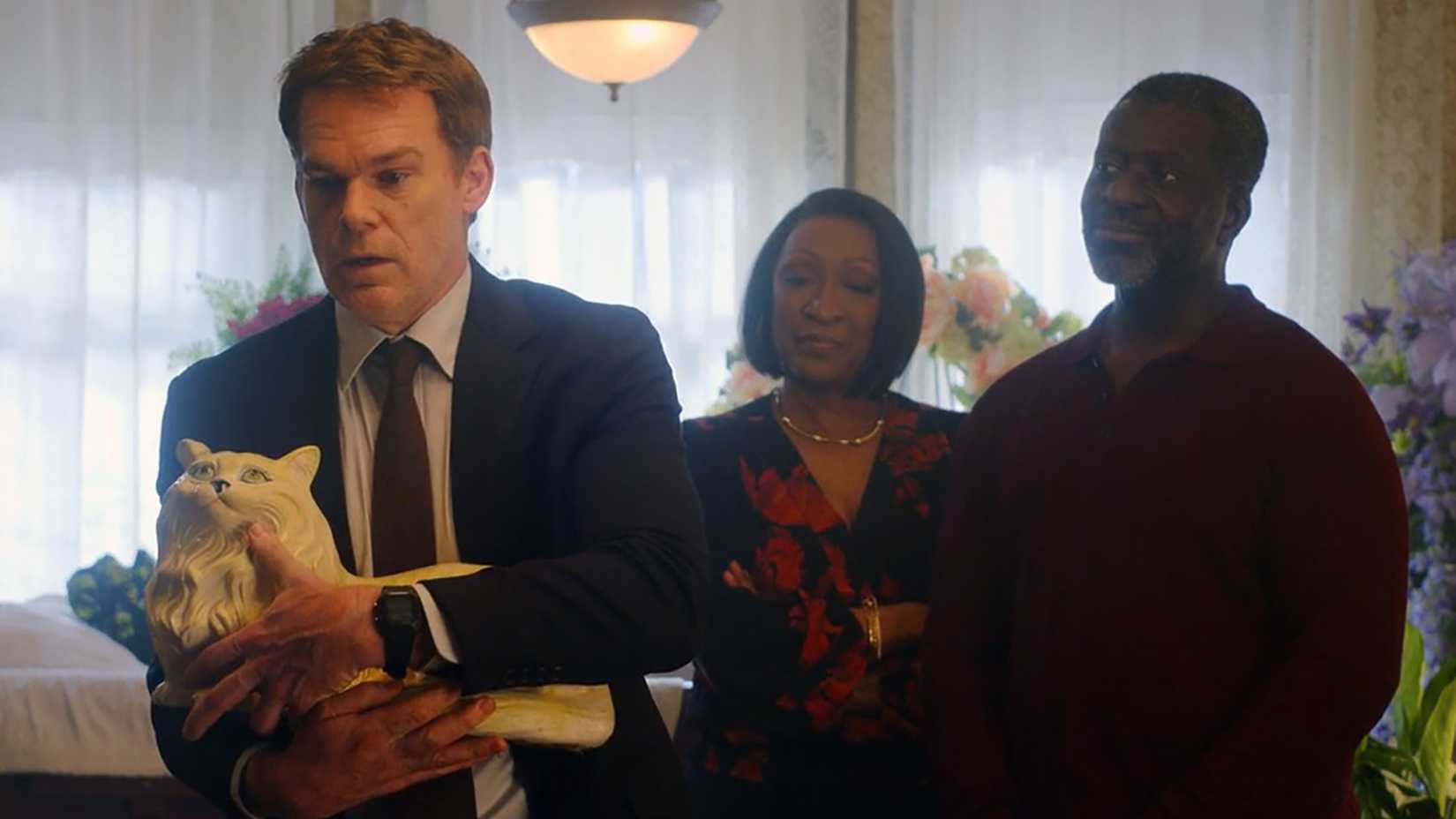 Dexter in a suit holding a large cat ornament as Blessing's family members look on in Dexter: Resurrection.